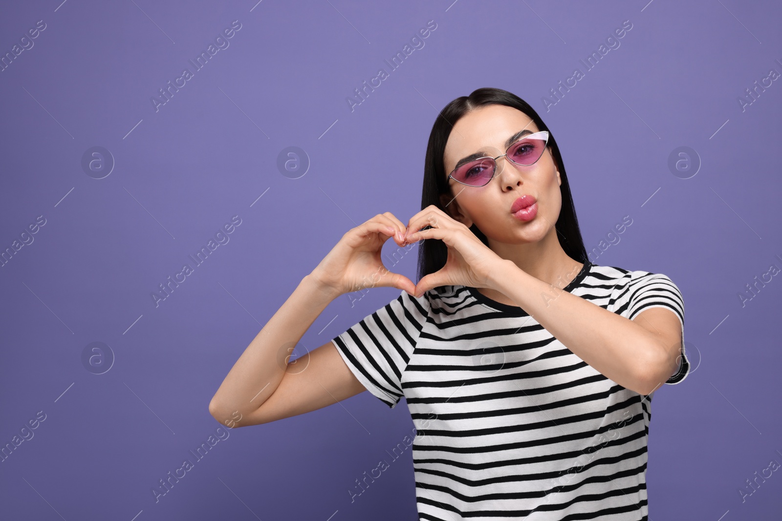 Photo of Beautiful young woman in stylish sunglasses blowing kiss and making heart with hands on purple background. Space for text