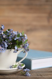 Photo of Beautiful forget-me-not flowers in cup, saucer and book on wooden table, closeup. Space for text