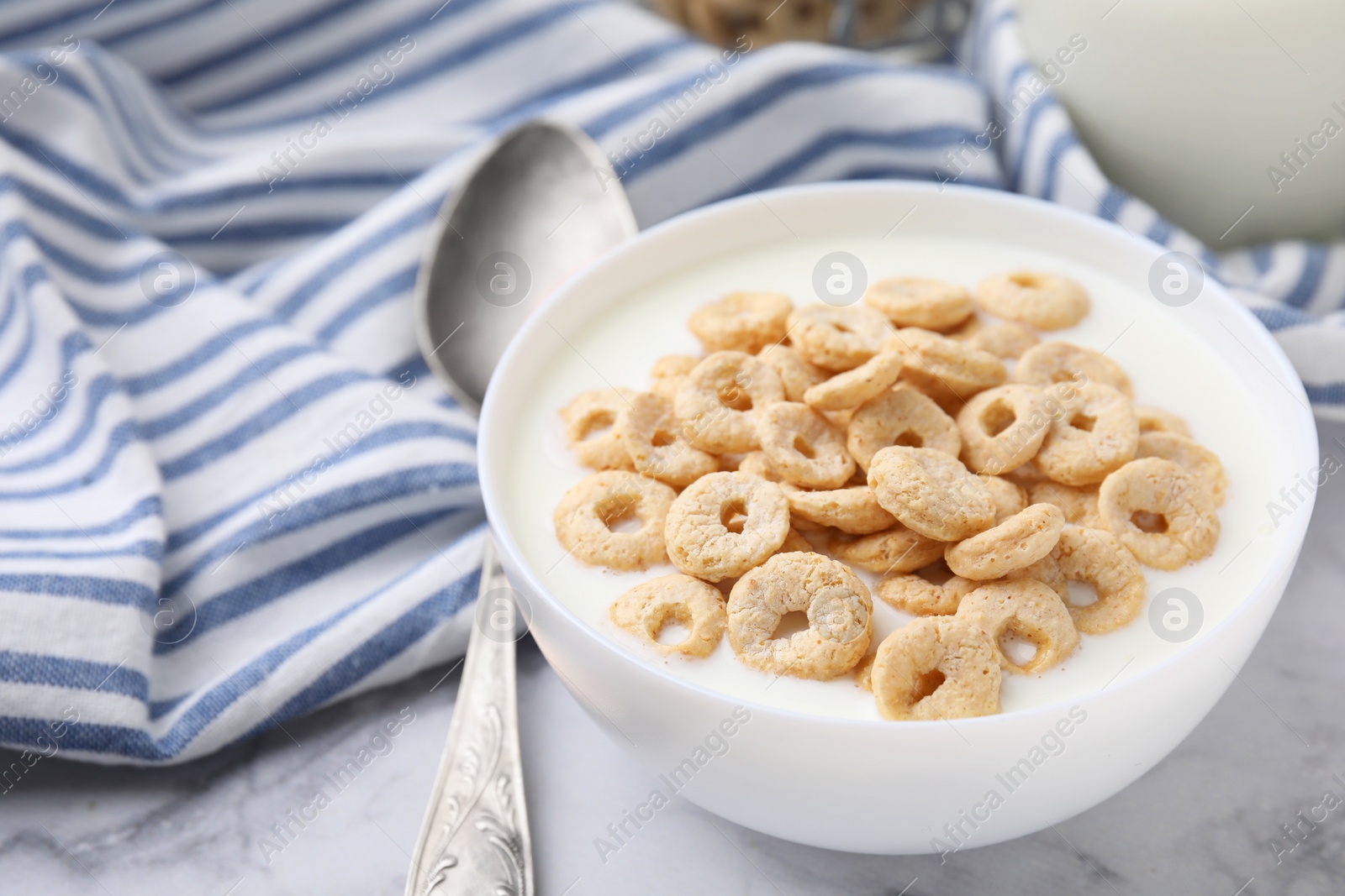 Photo of Breakfast cereal. Tasty corn rings with milk in bowl and spoon on white marble table, closeup