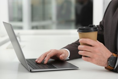 Photo of Man with cup of coffee working on laptop at white desk in office, closeup