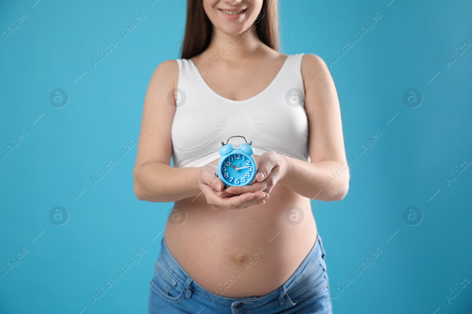 Photo of Young pregnant woman holding alarm clock near her belly on light blue background, closeup. Time to give birth