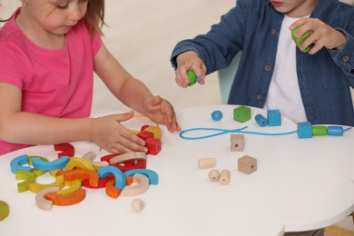 Photo of Little children playing with wooden pieces and string for threading activity at white table indoors, closeup. Developmental toys