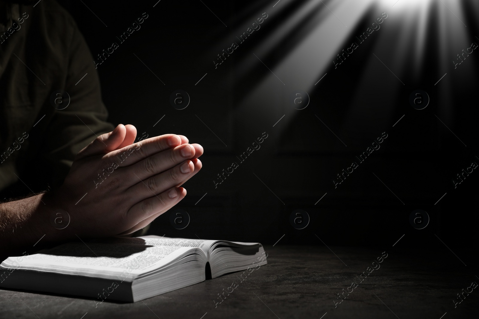 Image of Christian man praying over Bible in darkness, closeup. Holy light shining at him