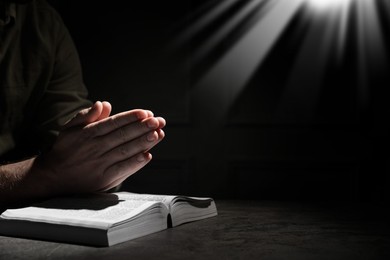 Image of Christian man praying over Bible in darkness, closeup. Holy light shining at him