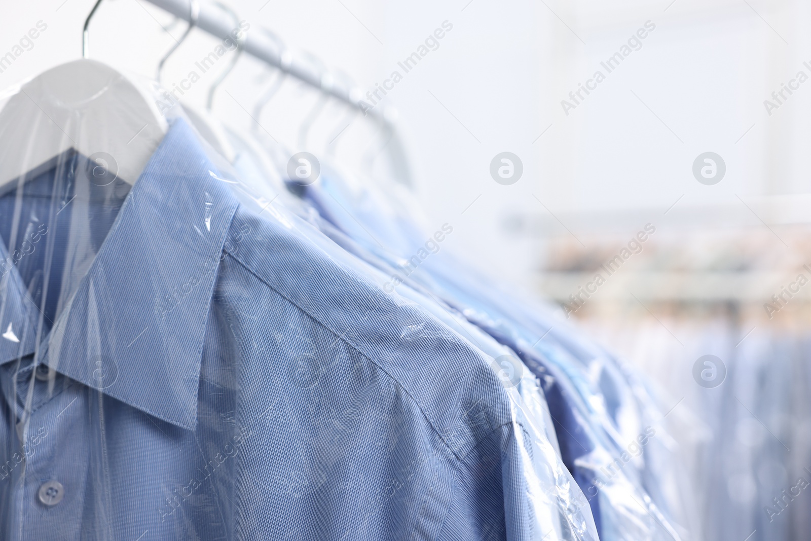 Photo of Dry-cleaning service. Many different clothes in plastic bags hanging on rack indoors, closeup