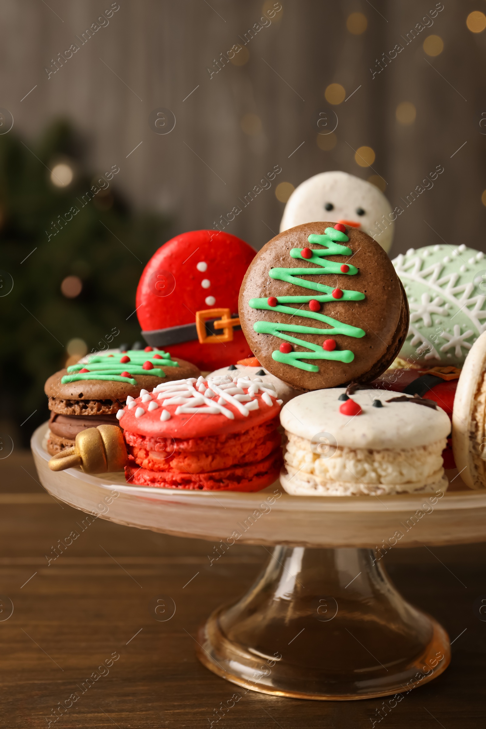 Photo of Stand with beautifully decorated Christmas macarons on wooden table against blurred festive lights