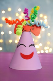 Photo of Funny handmade party hat on purple table against blurred lights