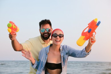 Happy couple with water guns near sea at sunset