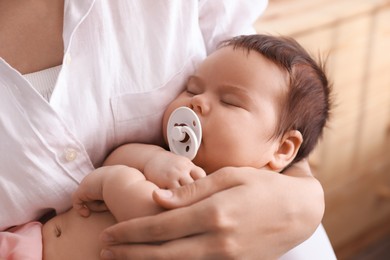 Mother holding her cute little baby with pacifier on blurred background, closeup