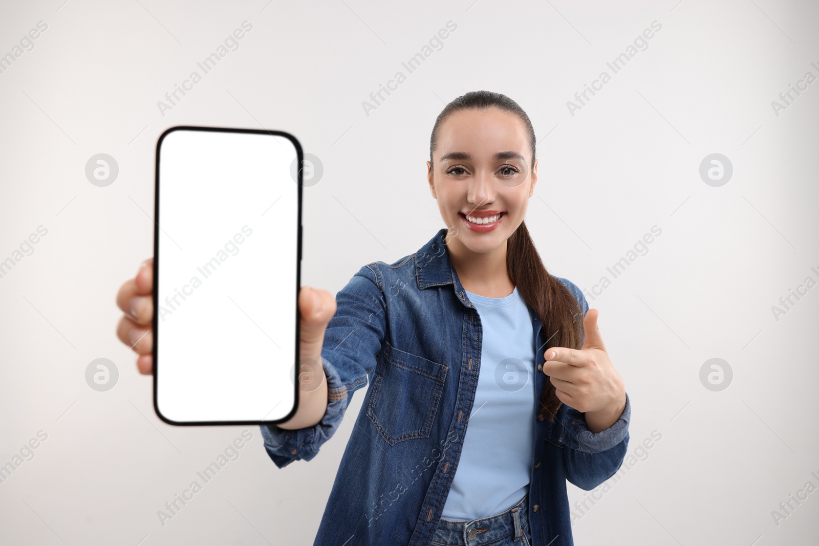 Photo of Young woman showing smartphone in hand and pointing at it on white background