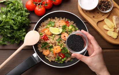 Photo of Woman adding sesame seeds to rice with shrimps and vegetables at wooden table, top view