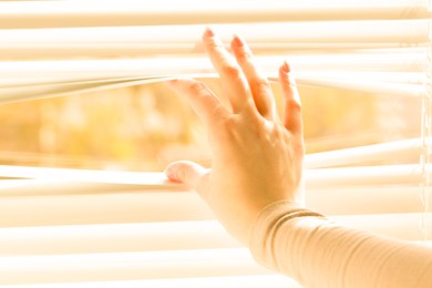 Woman separating slats of white blinds indoors, closeup