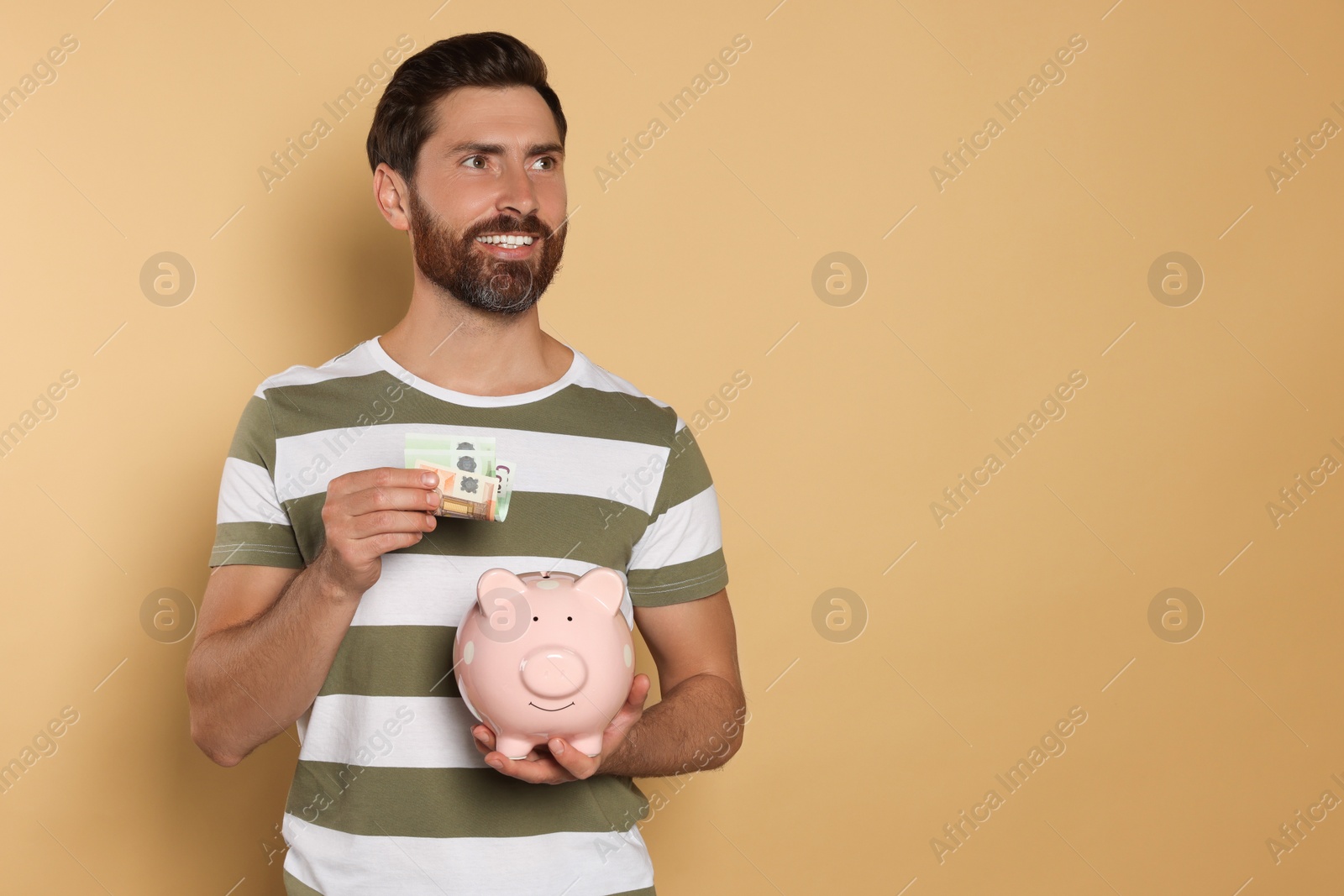 Photo of Happy man putting money into piggy bank on beige background, space for text