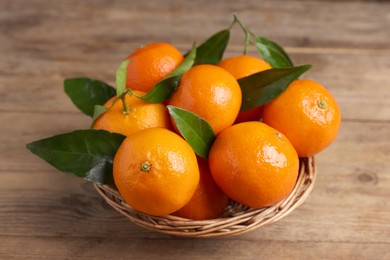 Delicious tangerines with leaves in wicker basket on wooden table, closeup