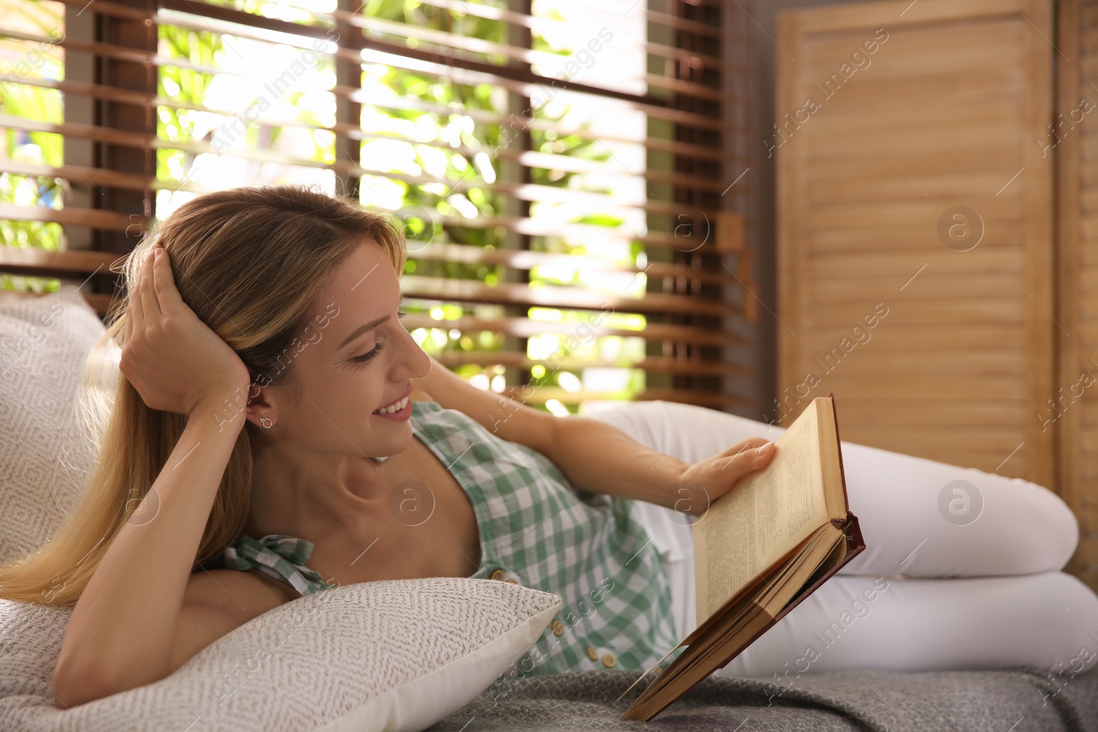 Photo of Beautiful young woman reading book near window at home