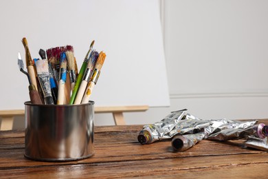 Photo of Easel with blank canvas and different art supplies on wooden table near white wall