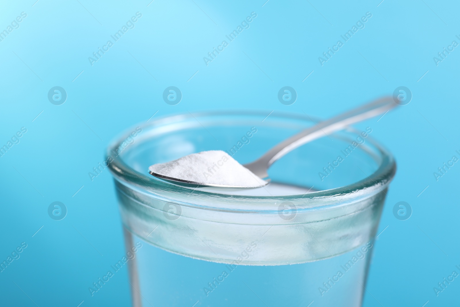 Photo of Glass of water and spoon with baking soda on light blue background, closeup