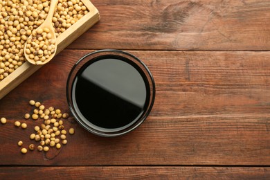 Photo of Soy sauce in bowl and soybeans on wooden table, flat lay. Space for text