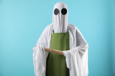 Woman in ghost costume and apron with rolling pin on light blue background