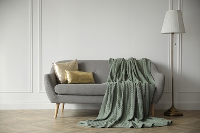 Photo of Stylish room interior with comfortable sofa, knitted blanket and lamp near white wall