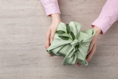 Photo of Furoshiki technique. Woman holding gift packed in green fabric and decorated with ruscus branch at wooden table, top view