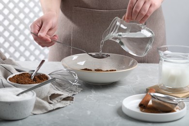 Photo of Making dalgona coffee. Woman pouring water into bowl at light gray table, closeup