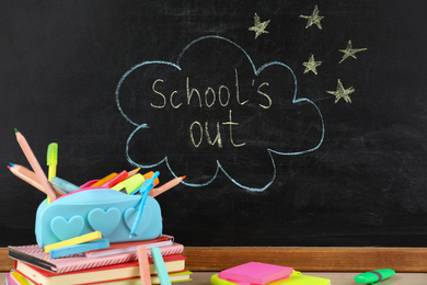 Photo of Pieces of color chalk and stationery on table near blackboard with text School's Out. Summer holidays