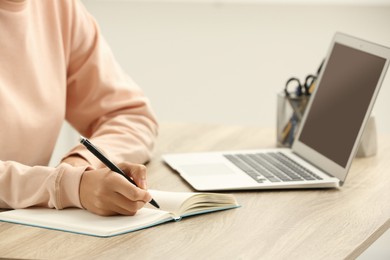 Photo of African American woman writing in notepad near laptop at wooden table indoors, closeup