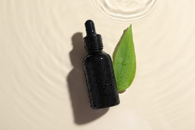 Bottle of cosmetic oil and green leaf in water on beige background, flat lay