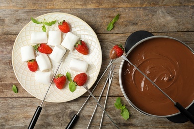 Flat lay composition with chocolate fondue in pot, strawberries and marshmallows on wooden background