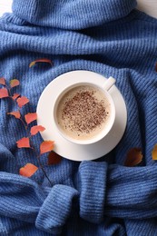 Cup of hot drink and leaves on blue knitted sweater, flat lay. Cozy autumn atmosphere