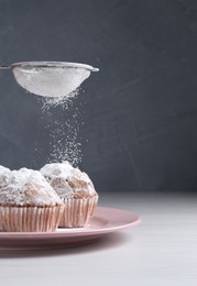 Woman with sieve sprinkling powdered sugar onto muffins at white wooden table, closeup