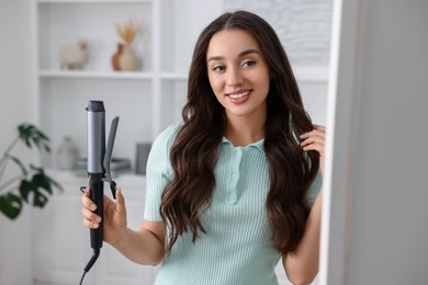 Smiling woman with beautiful hairstyle holding curling hair iron at home