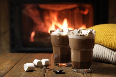 Photo of Glasses with hot cocoa, marshmallows and sweaters on wooden table near fireplace, space for text