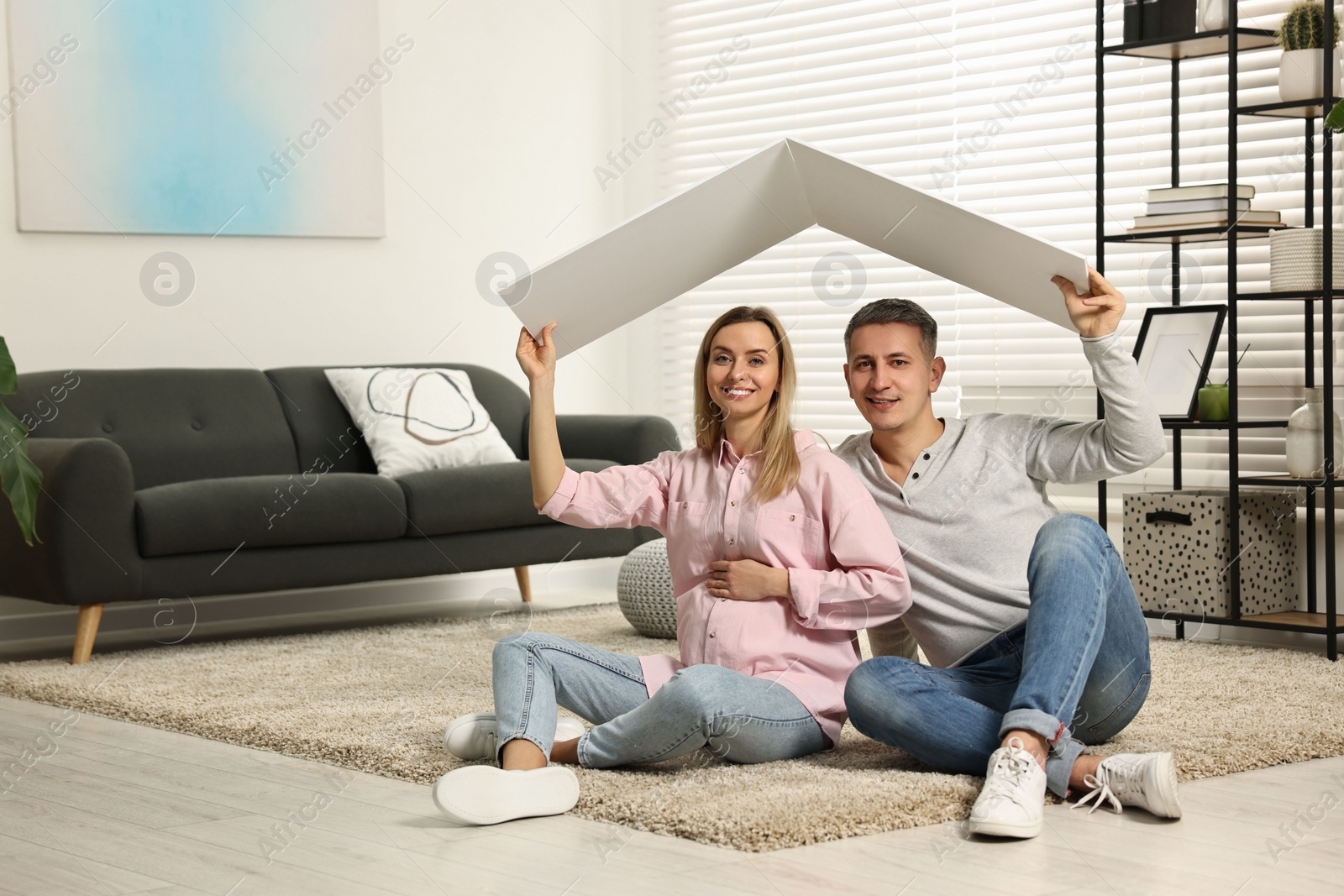 Photo of Young family housing concept. Pregnant woman with her husband sitting under cardboard roof on floor at home