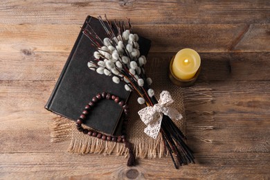 Rosary beads, Bible, burning candle and willow branches on wooden table, flat lay