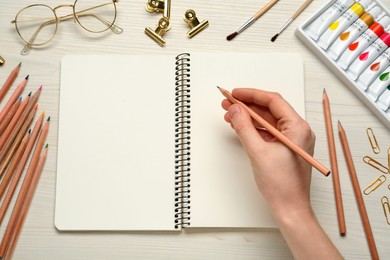 Woman with pencil and blank sketchbook at white wooden table, top view