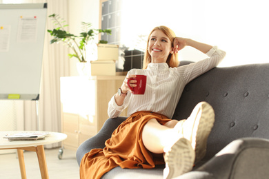 Young woman with cup of drink relaxing on couch at workplace