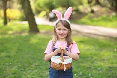Easter celebration. Cute little girl in bunny ears holding wicker basket with painted eggs outdoors