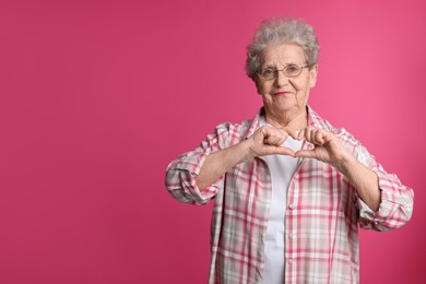 Photo of Elderly woman making heart with her hands on pink background, space for text