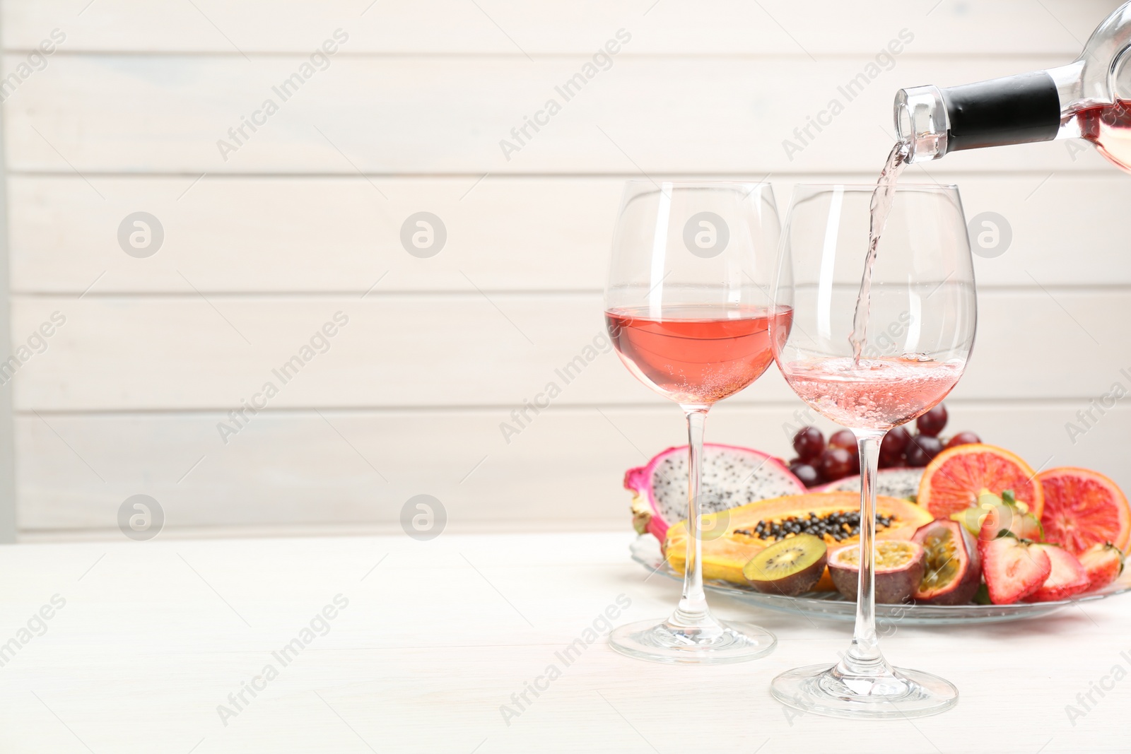 Photo of Pouring wine into glasses and delicious exotic fruits on white wooden table. Space for text