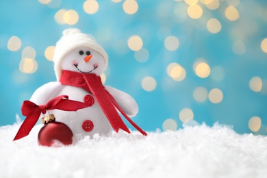 Photo of Snowman toy and Christmas ball on snow against blurred festive lights. Space for text