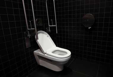 Photo of Clean ceramic toilet bowl and handrails on tiled wall indoors