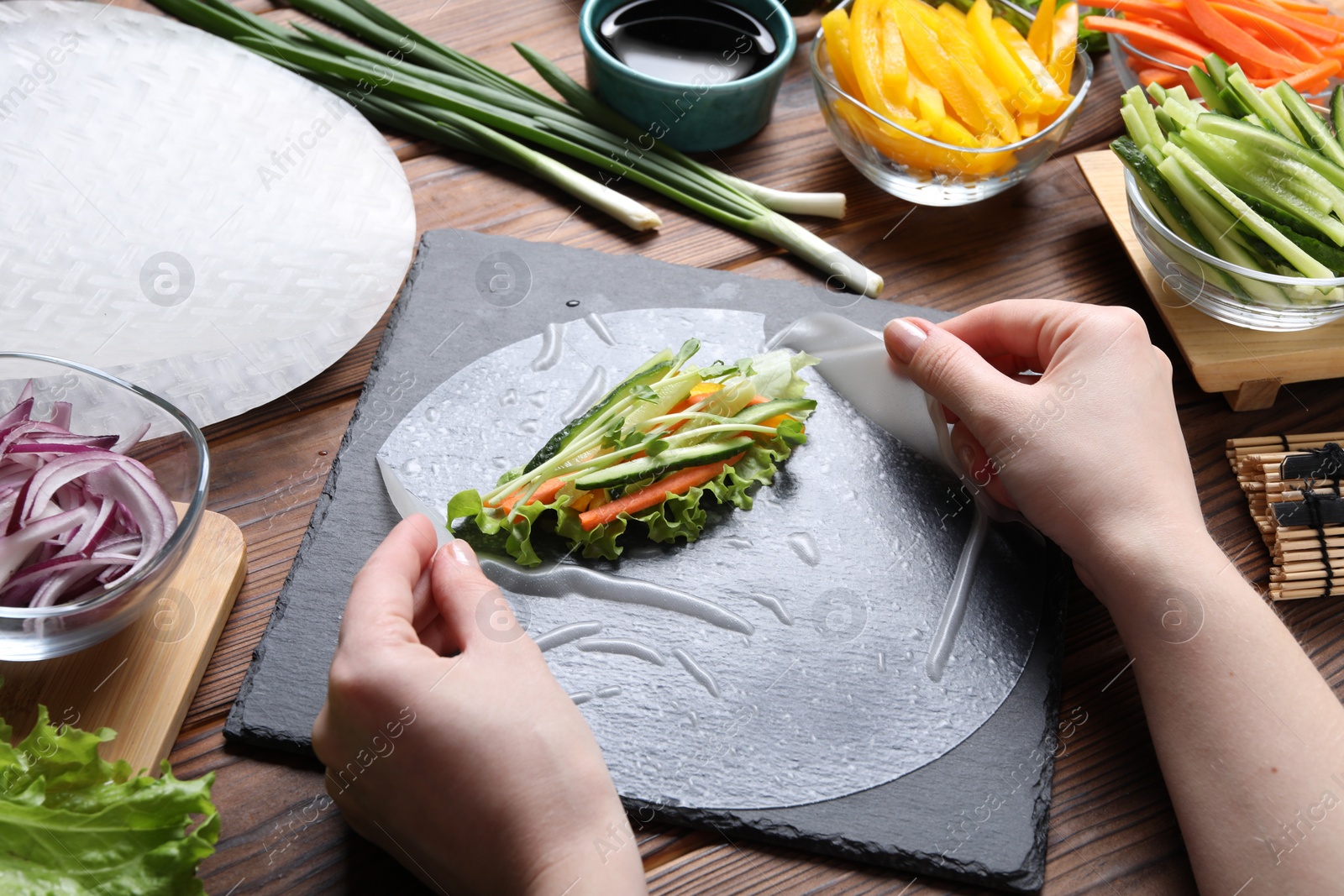 Photo of Woman wrapping spring roll at wooden table with products, closeup