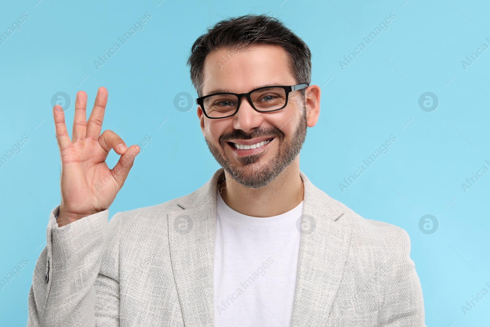 Photo of Portrait of happy man in stylish glasses showing OK gesture on light blue background