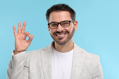 Photo of Portrait of happy man in stylish glasses showing OK gesture on light blue background