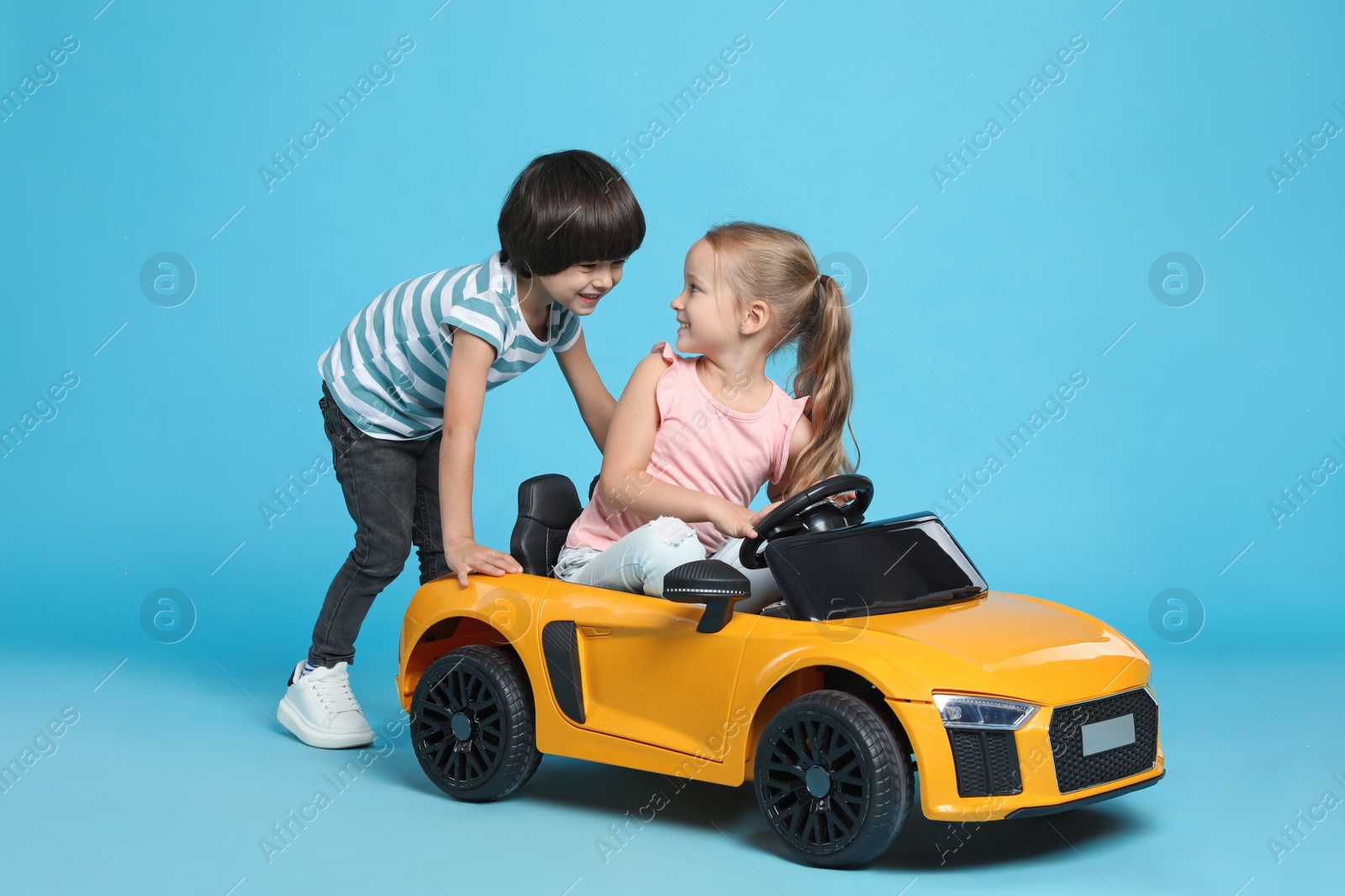 Photo of Cute boy pushing children's electric toy car with little girl on light blue background