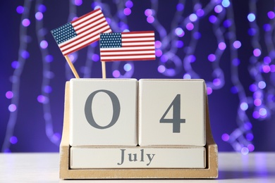 Photo of Wooden calendar with USA flags on table against blurred lights, closeup. Happy Independence Day
