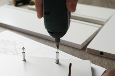 Photo of Man with electric screwdriver assembling white furniture at table, closeup