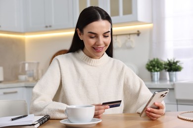 Happy young woman with smartphone and credit card shopping online at wooden table in kitchen